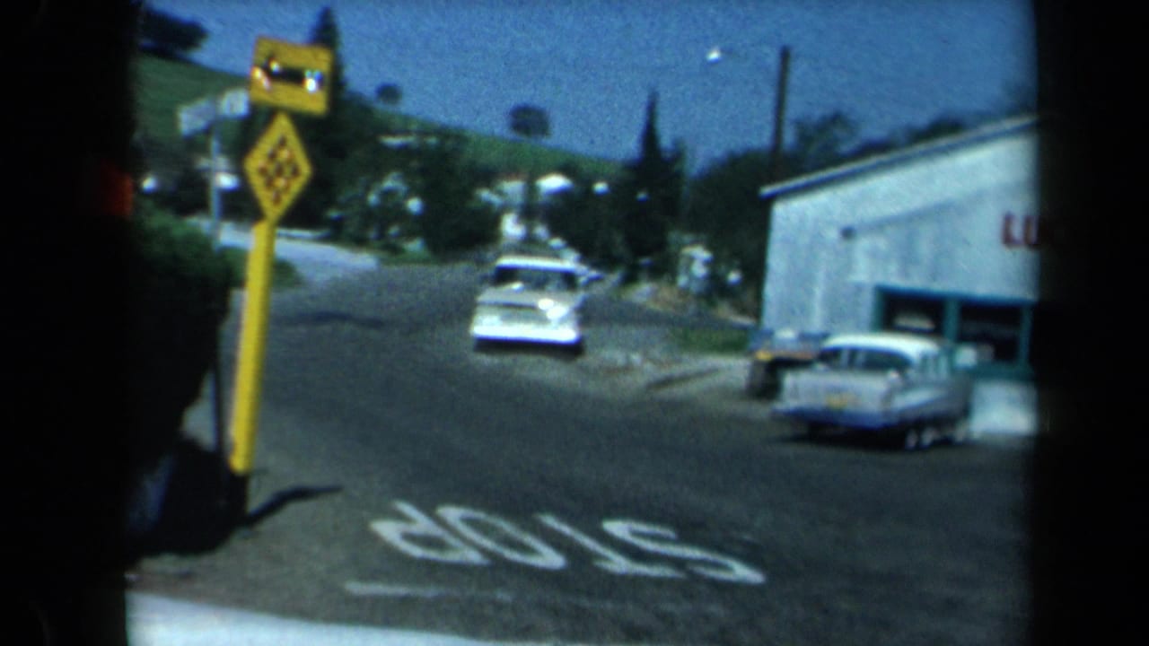 1969 California 8mm frame showing a rural roadside stop, white sedan, yellow warning sign, STOP pavement marking, and small store building.