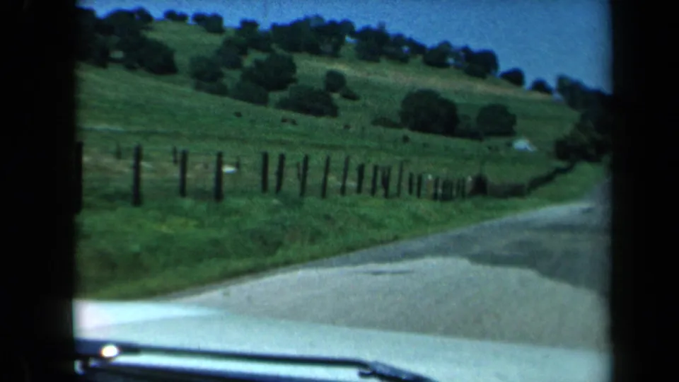 1969 California road-trip view from inside a car, looking toward a curving rural road, green hillside, fence line, and scattered trees.