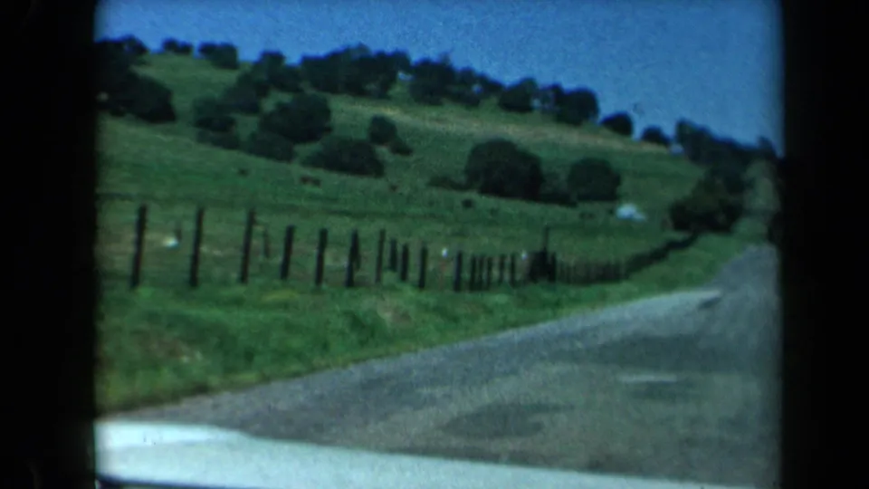 1969 California 8mm travel frame showing a bend in a country road beside fence posts, green pasture, trees, and a car hood edge.