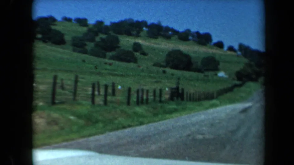1969 California home-movie frame of a winding rural road beside wooden fencing, rolling green pasture, and scattered trees.