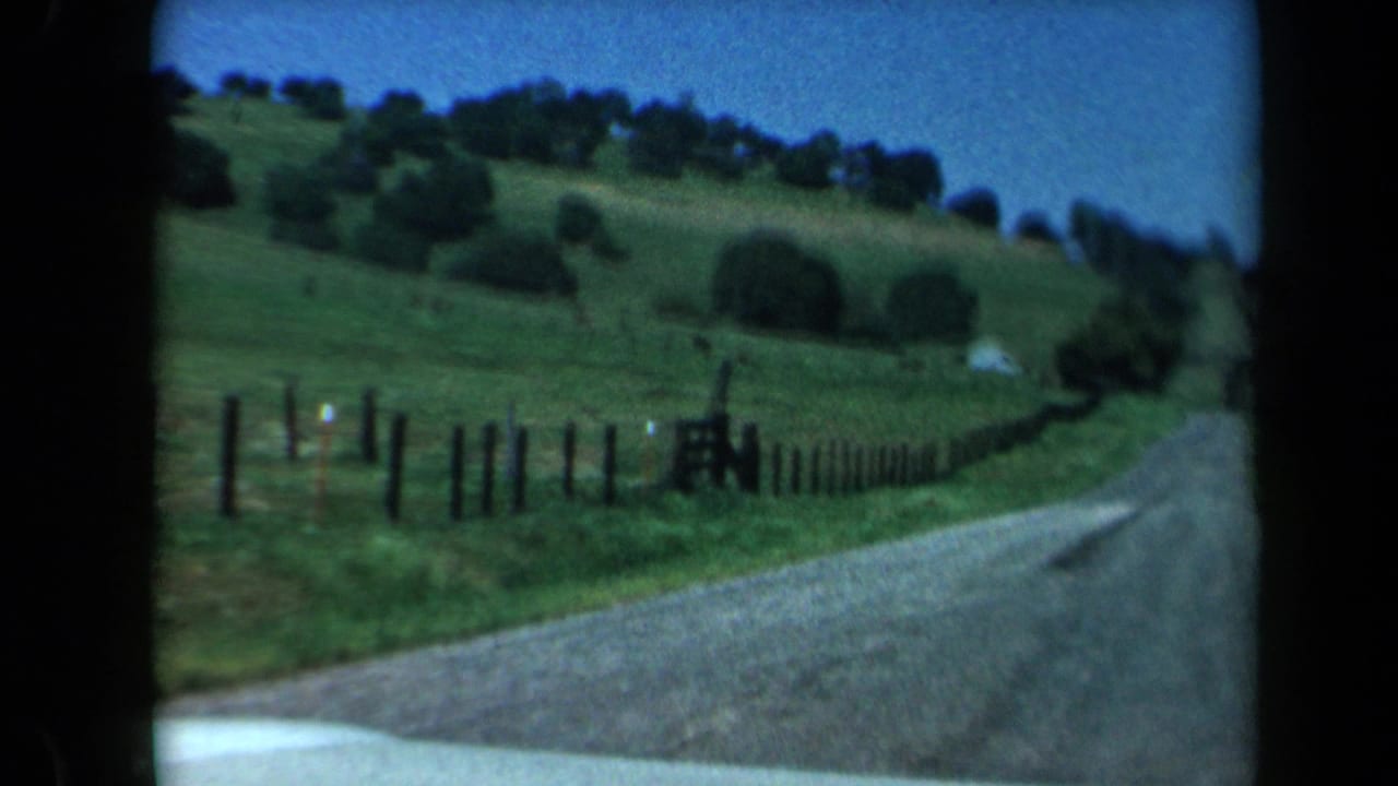 1969 California home-movie frame showing a road curving beside green hills, trees, and wooden fencing from a moving car.
