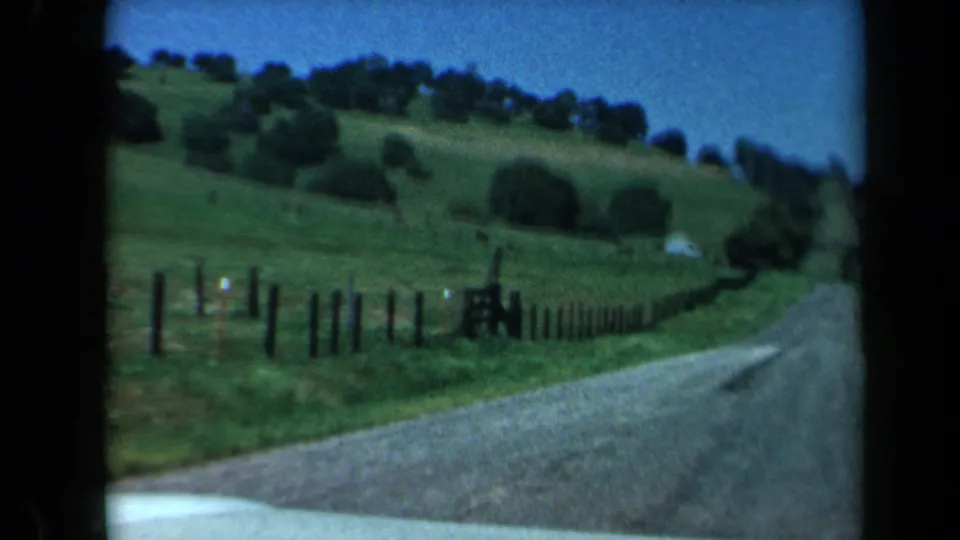 1969 California home-movie frame showing a road curving beside green hills, trees, and wooden fencing from a moving car.