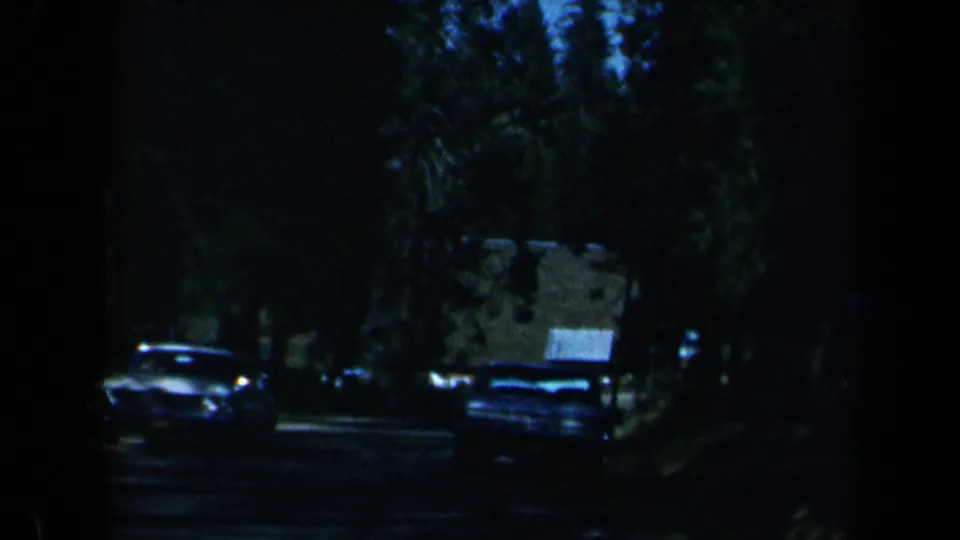 1969 California home-movie frame of a dark wooded roadside with parked American cars and a partly visible building.