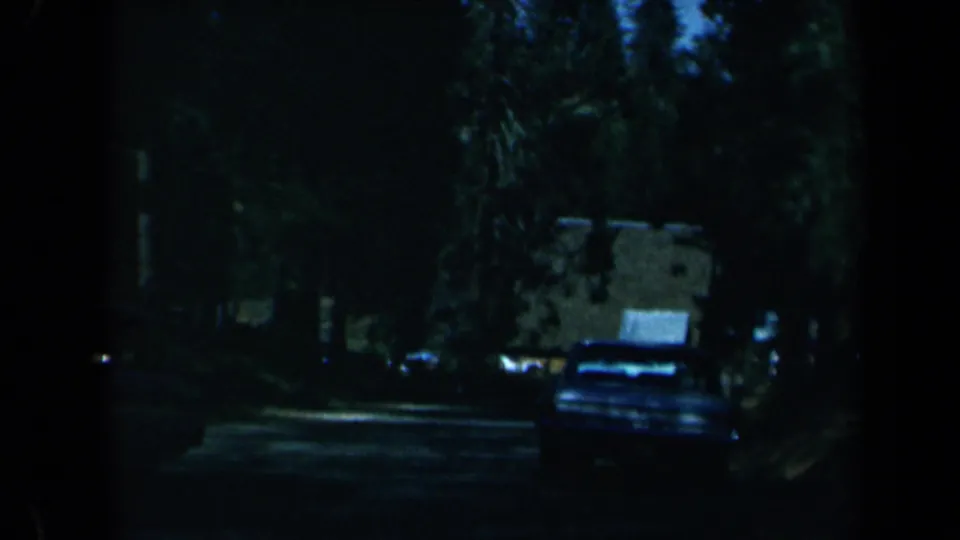 1969 California wooded road scene with a parked car, dense trees, and a small roadside building in low light.