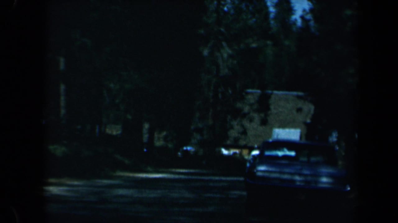 1969 California 8mm frame showing a shadowed tree-lined road, a classic car ahead, and low roadside buildings in the distance.