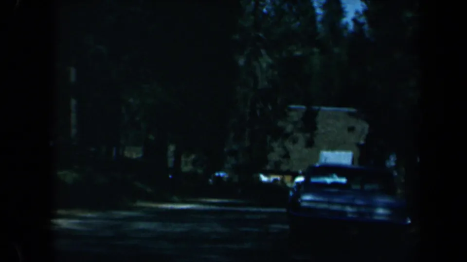 1969 California 8mm frame showing a shadowed tree-lined road, a classic car ahead, and low roadside buildings in the distance.