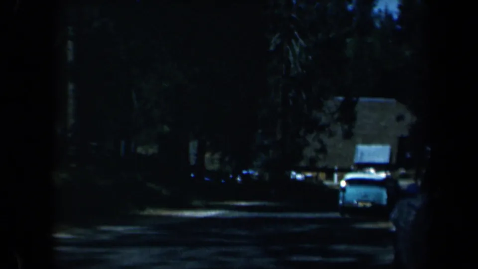 1969 California 8mm frame with a light-colored car on a dark wooded street and low structures set back behind trees.