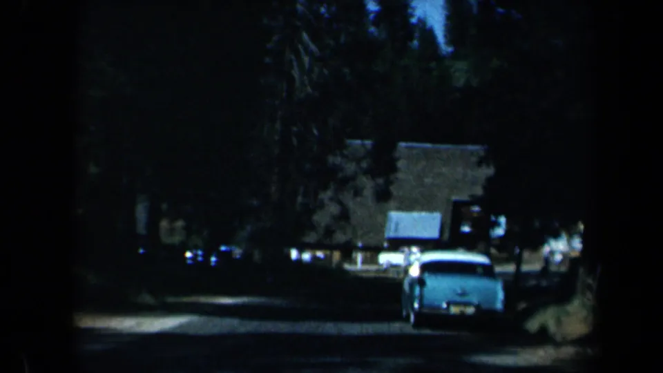 1969 California 8mm frame showing the rear of a classic car on a shaded road near trees, parked cars, and low buildings.