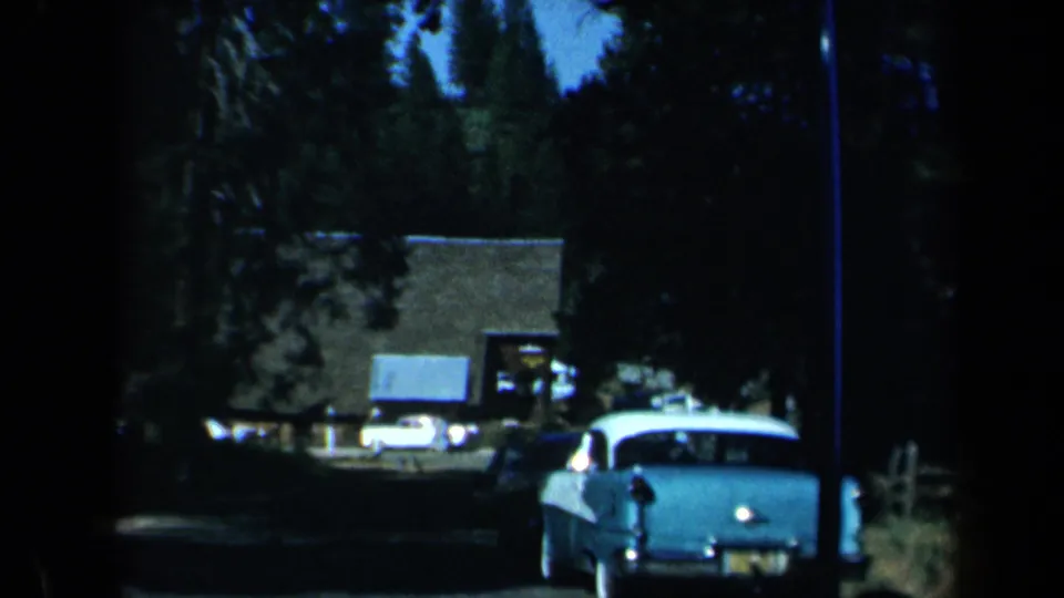1969 California 8mm frame showing a light classic car from behind on a tree-lined road with buildings and parked cars in the distance.