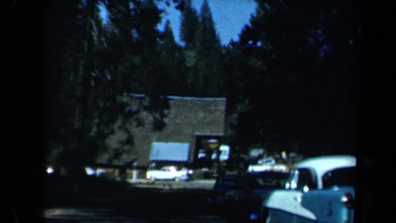 1969 California roadside scene with parked cars, a rustic structure, trees, and dark foreground framing from a vehicle or shaded position.