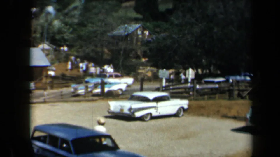 1969 California gravel lot with classic American cars, wooden fencing, visitors in light clothing, trees, and rustic structures in the background.