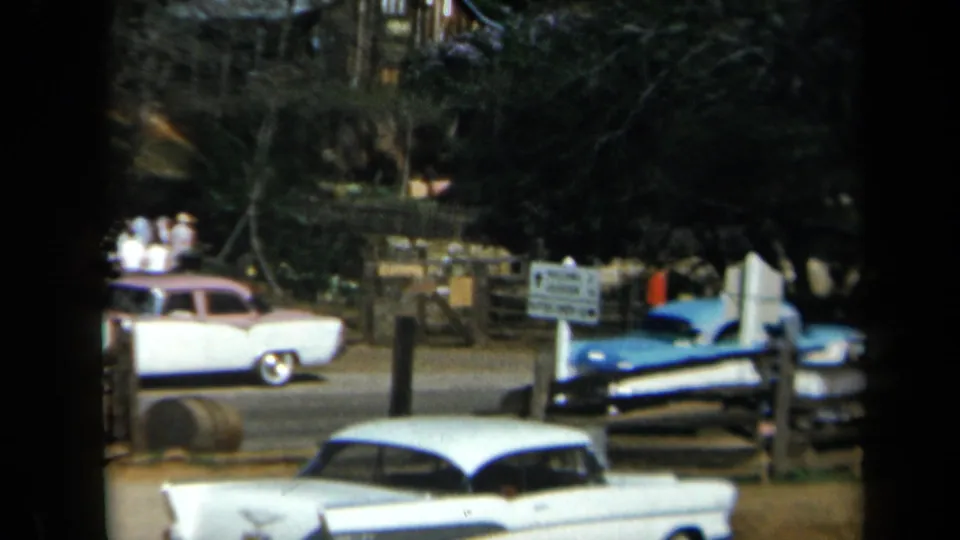 1969 California roadside stop with parked and passing cars, a wooden fence, posted sign, visitors, and trees behind the lot.