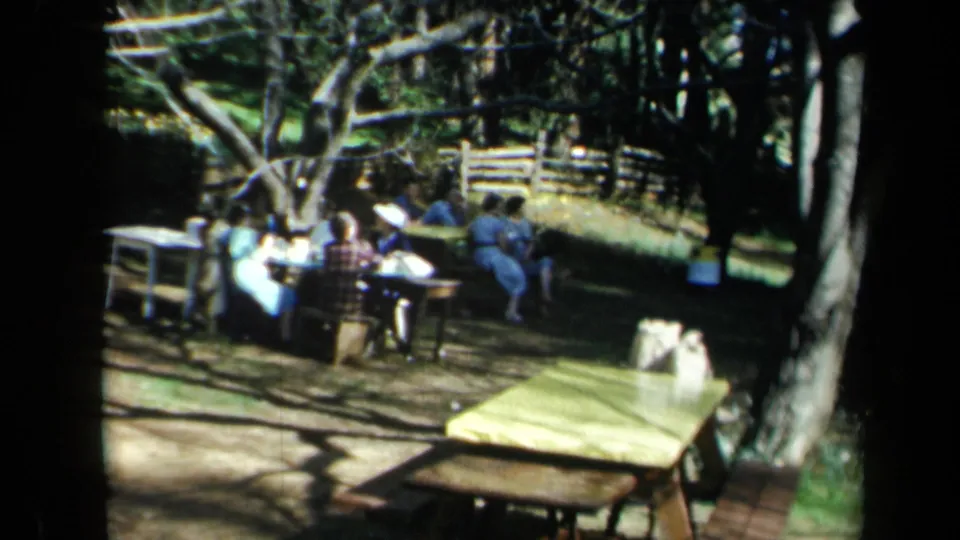 Adults in period clothing sit around picnic tables in a wooded California park, with fencing and open ground behind them.