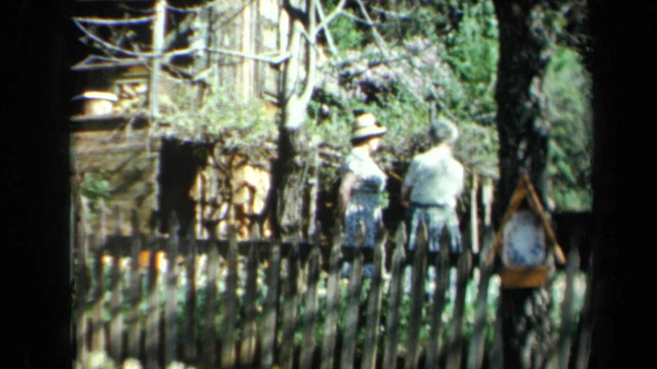 1969 California 8mm frame showing adults by a picket fence beside blooming lilacs and weathered wood siding.