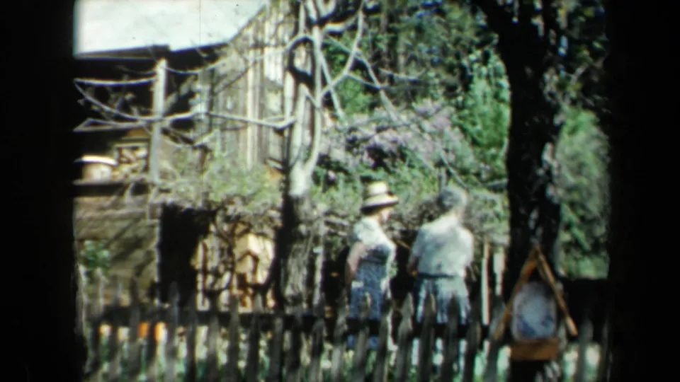 1969 California home-movie frame with adults near a wooden fence, purple lilacs, trees, and rustic yard details.