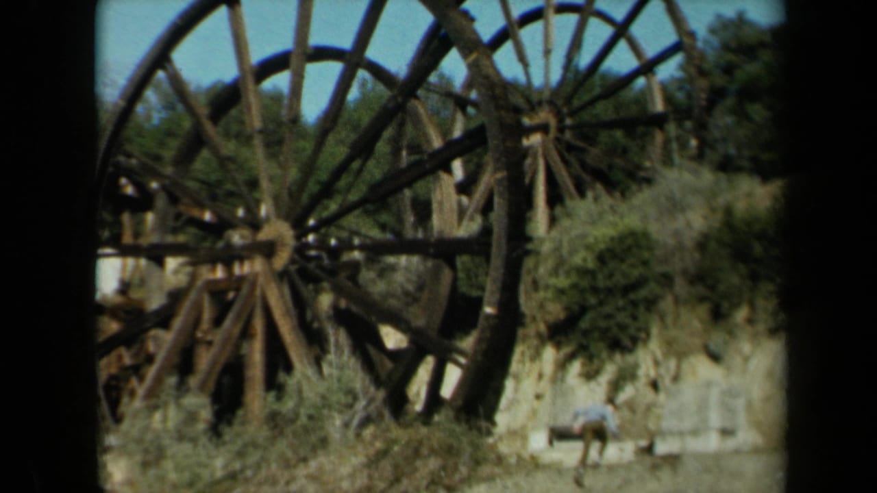 1969 California 8mm frame showing oversized weathered wooden wheels, dry brush, trees, blue sky, and a distant person below.