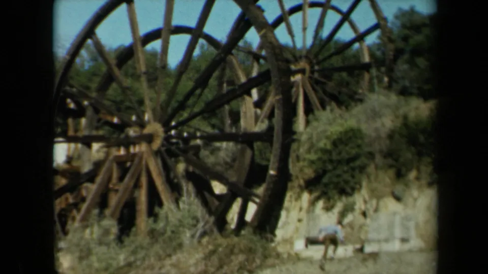 1969 California 8mm frame showing oversized weathered wooden wheels, dry brush, trees, blue sky, and a distant person below.