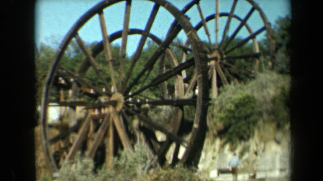 Two large wooden wheels fill the frame with brush, trees, sky, and a small figure below.