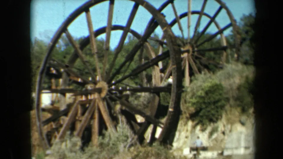 1969 California 8mm frame showing a close view of a weathered wooden wheel and a distant visitor near the lower edge.