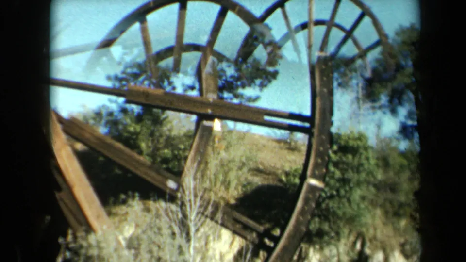1969 California home-movie frame looking through the wheel structure at angled beams, vegetation, trees, and blue sky.