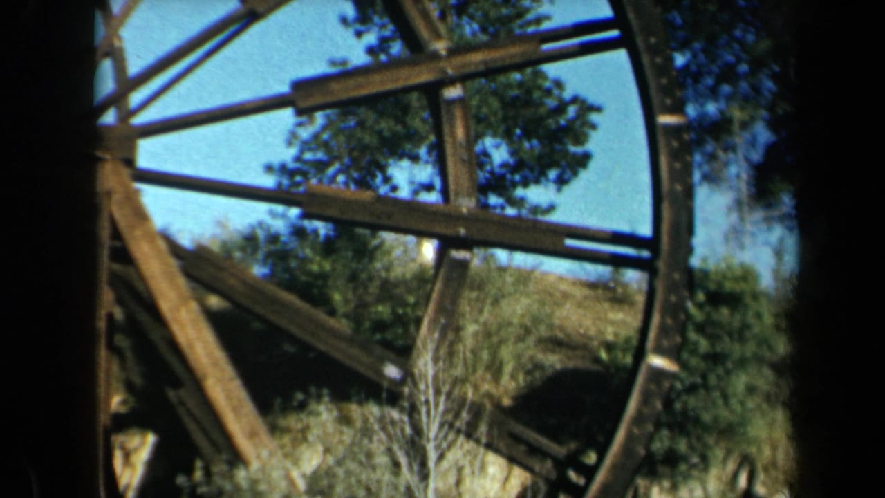 1969 California 8mm frame showing a large weathered wooden waterwheel with beams, spokes, trees, and brush nearby.