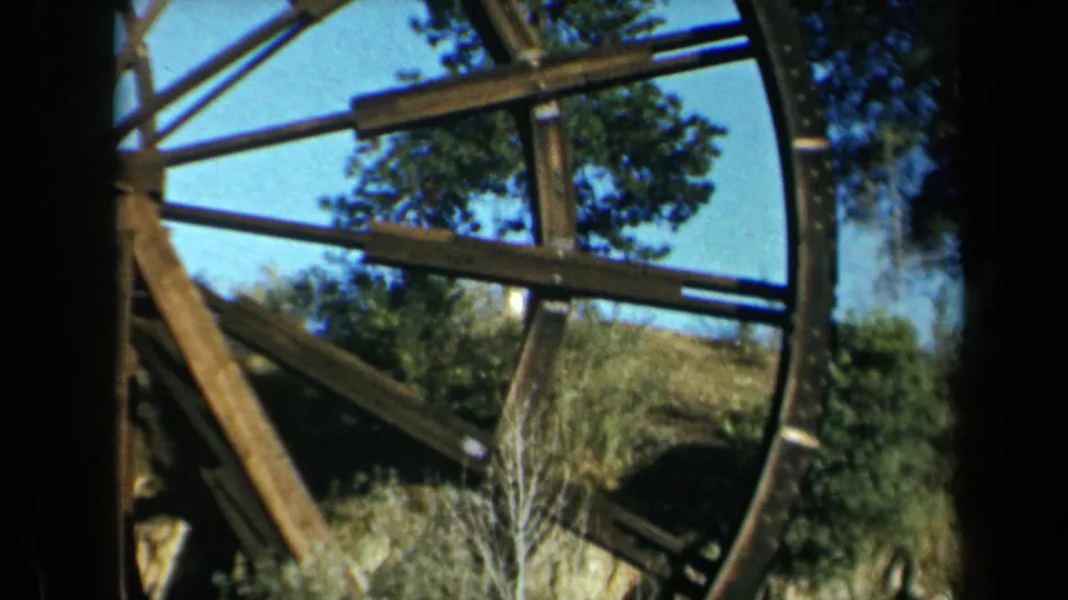 1969 California 8mm frame showing a large weathered wooden waterwheel with beams, spokes, trees, and brush nearby.