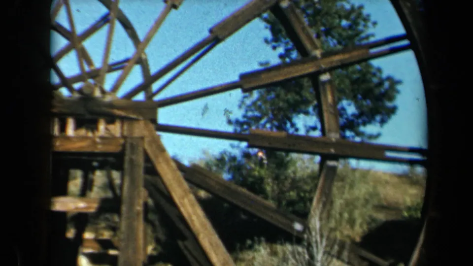 1969 California 8mm frame of the waterwheel's central wooden frame, beams, spokes, trees, and bright sky.