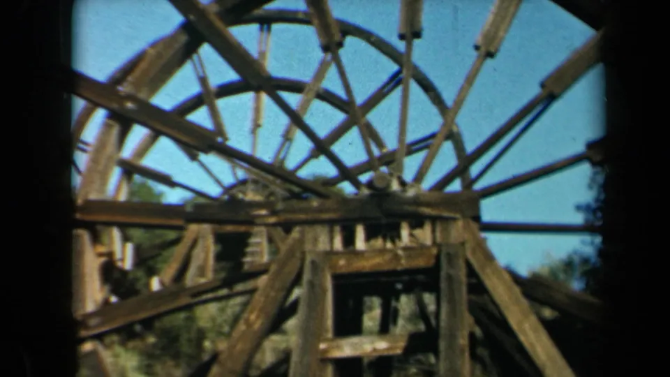 1969 California 8mm frame showing the waterwheel framework with a distant adult visitor partly visible beside the beams.