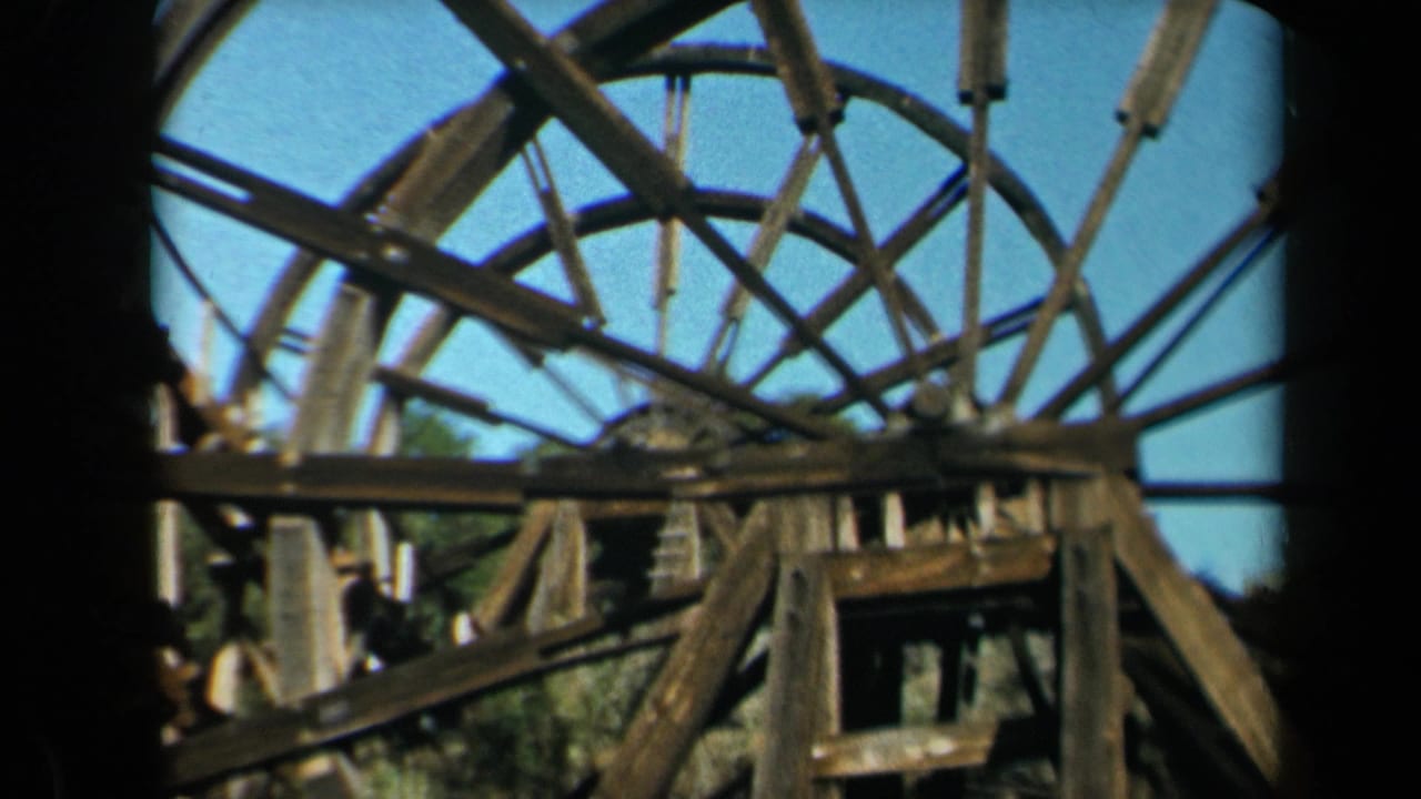 1969 California 8mm frame showing a weathered wooden spoke waterwheel and timber support beams under a blue sky.