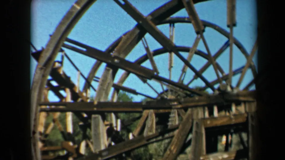 A vintage 8mm view of an old wooden waterwheel structure in California, with spokes and beams silhouetted against daylight sky.