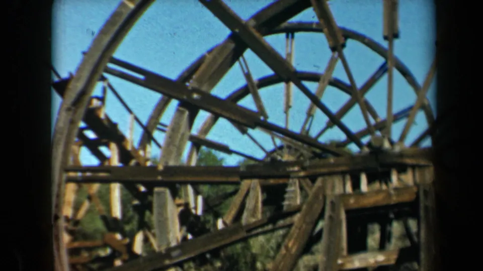 1969 home-movie frame showing the rim and spokes of a weathered wooden waterwheel, partly framed by dark 8mm borders.