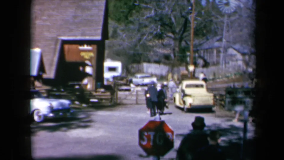 A California mountain-town street scene shows pedestrians, classic cars, a stop sign, and a wooden building in 1969 home-movie footage.