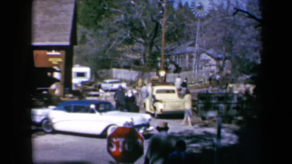 A white mid-century car crosses the foreground as pedestrians gather near parked vehicles on a 1969 California town street.