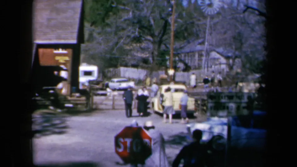 People walk and gather near classic cars, fencing, bare trees, and a stop sign in 1969 California 8mm street footage.