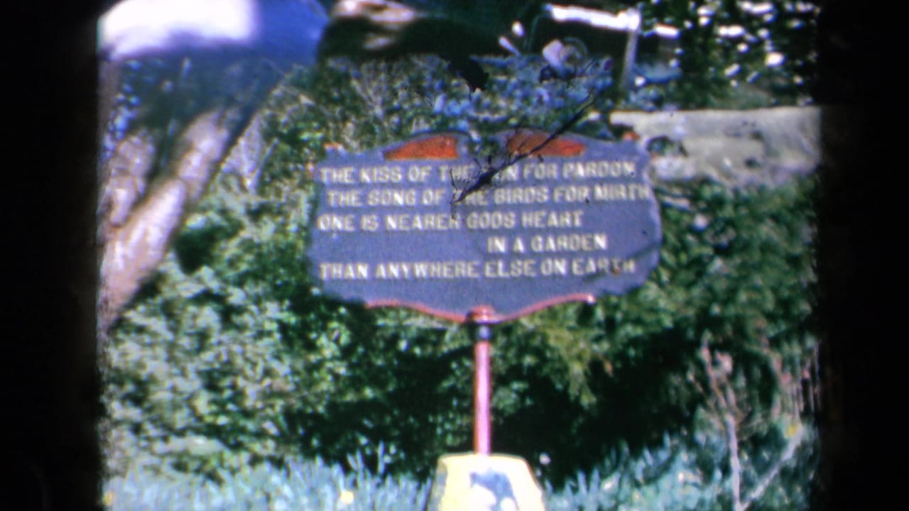 A garden sign with white verse lettering stands among greenery, partly obscured by film-frame edges.