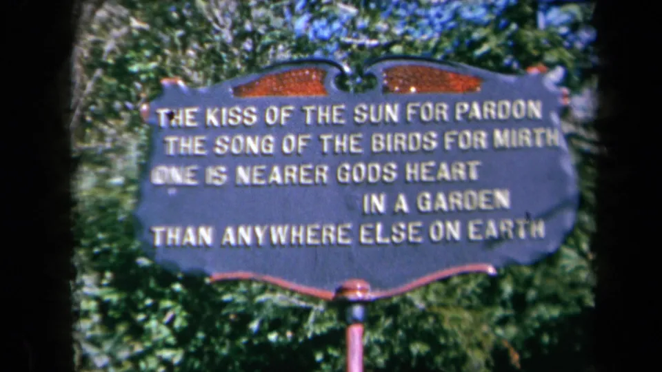 Close 1969 California home-movie view of a scalloped metal garden sign bearing a poetic verse, surrounded by greenery.