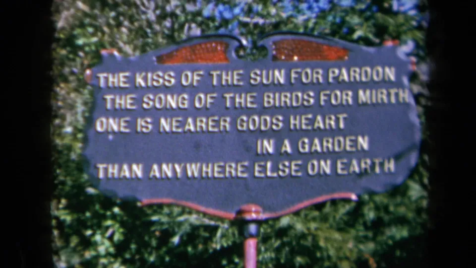 Final 1969 California frame of the decorative garden verse sign, centered in a handheld 8mm view with foliage behind it.