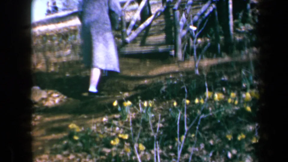 In 1969 California home-movie footage, a coat-wearing person climbs rustic wooden steps past daffodils and shrubs.