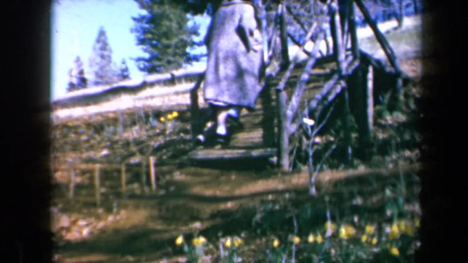 A person in a long coat and hat moves up a rustic garden stairway in California, with yellow daffodils nearby.