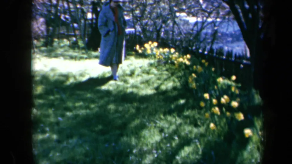 In California 1969 home-movie footage, a woman in a long coat walks through a shaded garden beside yellow daffodils.