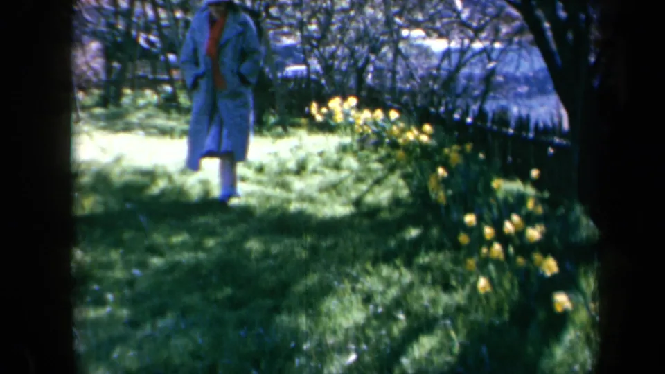 A woman in a blue coat and red scarf appears near daffodils in a shaded California garden, filmed on 8mm in 1969.