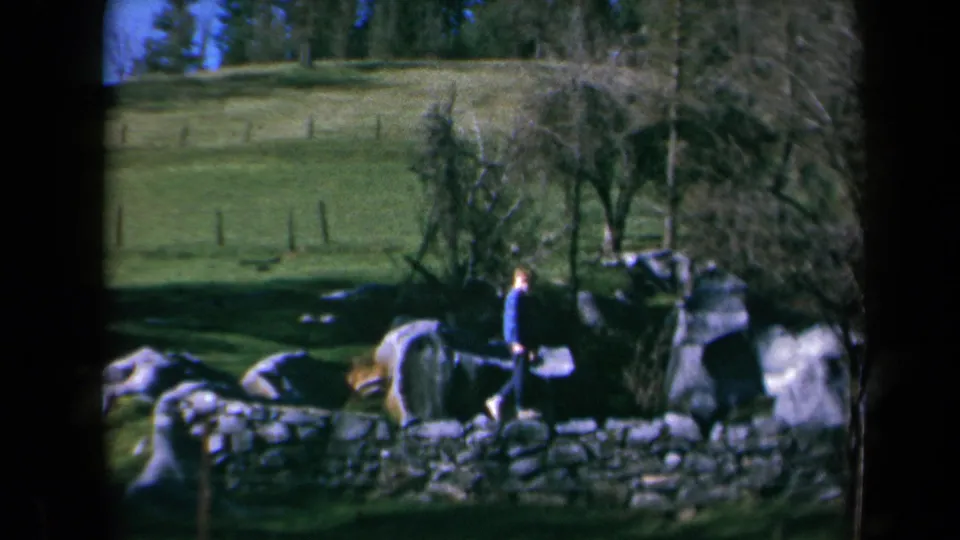 1969 California 8mm frame of a boy in a blue jacket walking past large rocks, a low stone wall, and leafless trees in a green pasture.