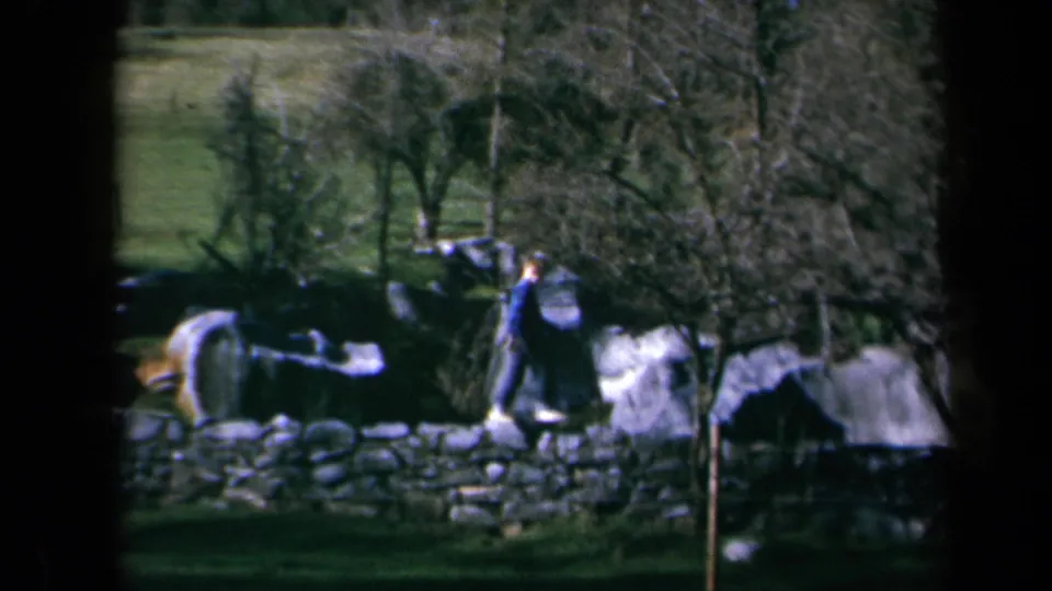 1969 California 8mm frame showing a boy in a blue jacket near a stone wall, large boulders, bare trees, and green hillside pasture.