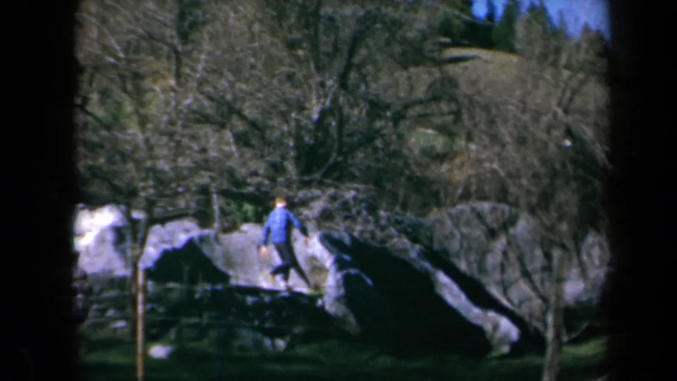 1969 California home-movie frame of a child moving off a rock in a wooded landscape with bare trees and grass.