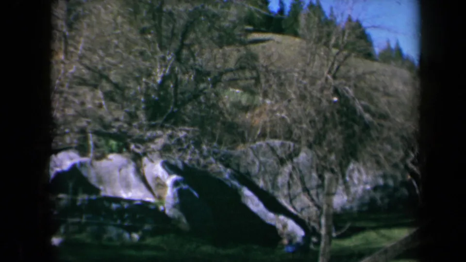 1969 California home-movie frame with the child low near the grass after the rock jump, surrounded by trees and hillside.