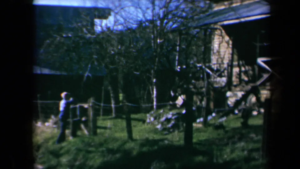 The pan holds on the boy near the fence as a weathered rural building and leafless trees fill the background.