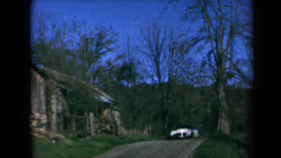 1969 California 8mm frame showing a white classic sedan approaching on a dirt road near a rustic cabin and trees.