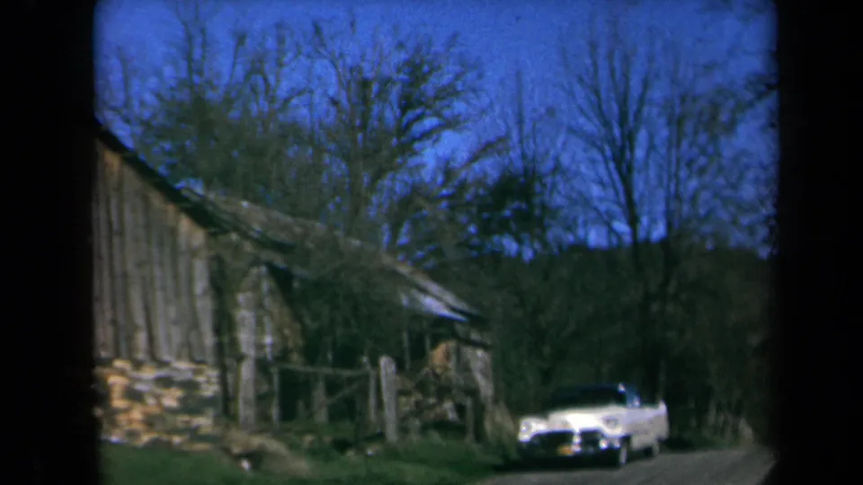 1969 California home-movie frame of a white sedan beside the rustic cabin, fence posts, stonework, trees, and dirt road.