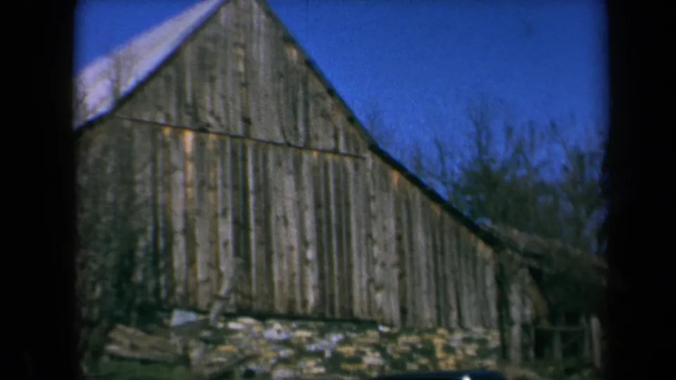 1969 California 8mm frame of a sloped-roof wooden barn with a stone foundation and trees behind it.