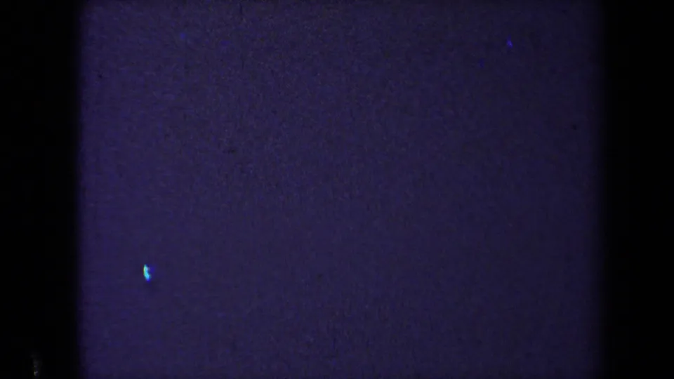 Purple-tinted 8mm film passage with little visible scene detail, showing grain, darkness, and a small blue mark.